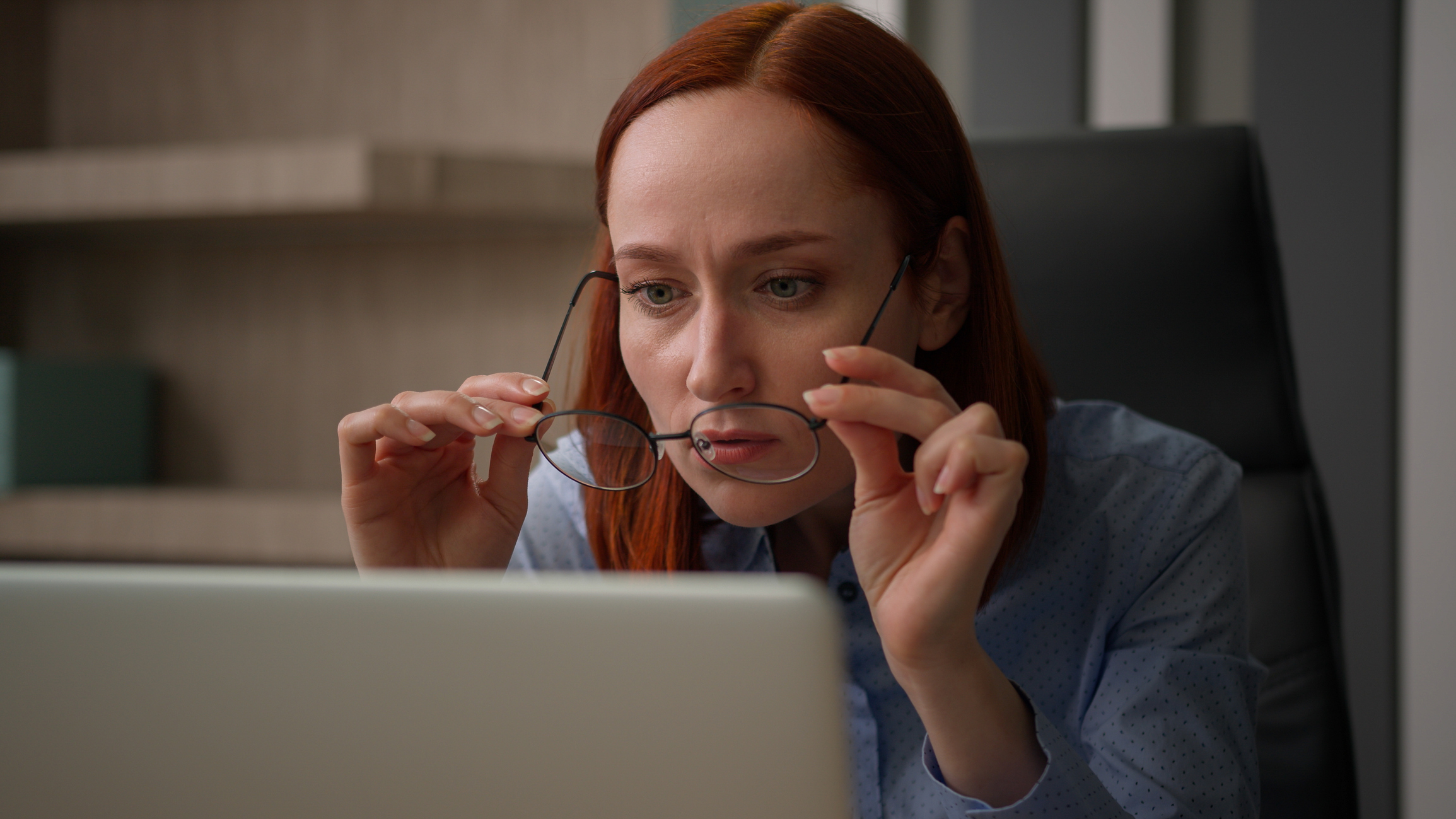 A Caucasian businesswoman working on a laptop in an office setting removes her glasses to inspect something on screen that is... problematic for ai-generated-google-ads blog.