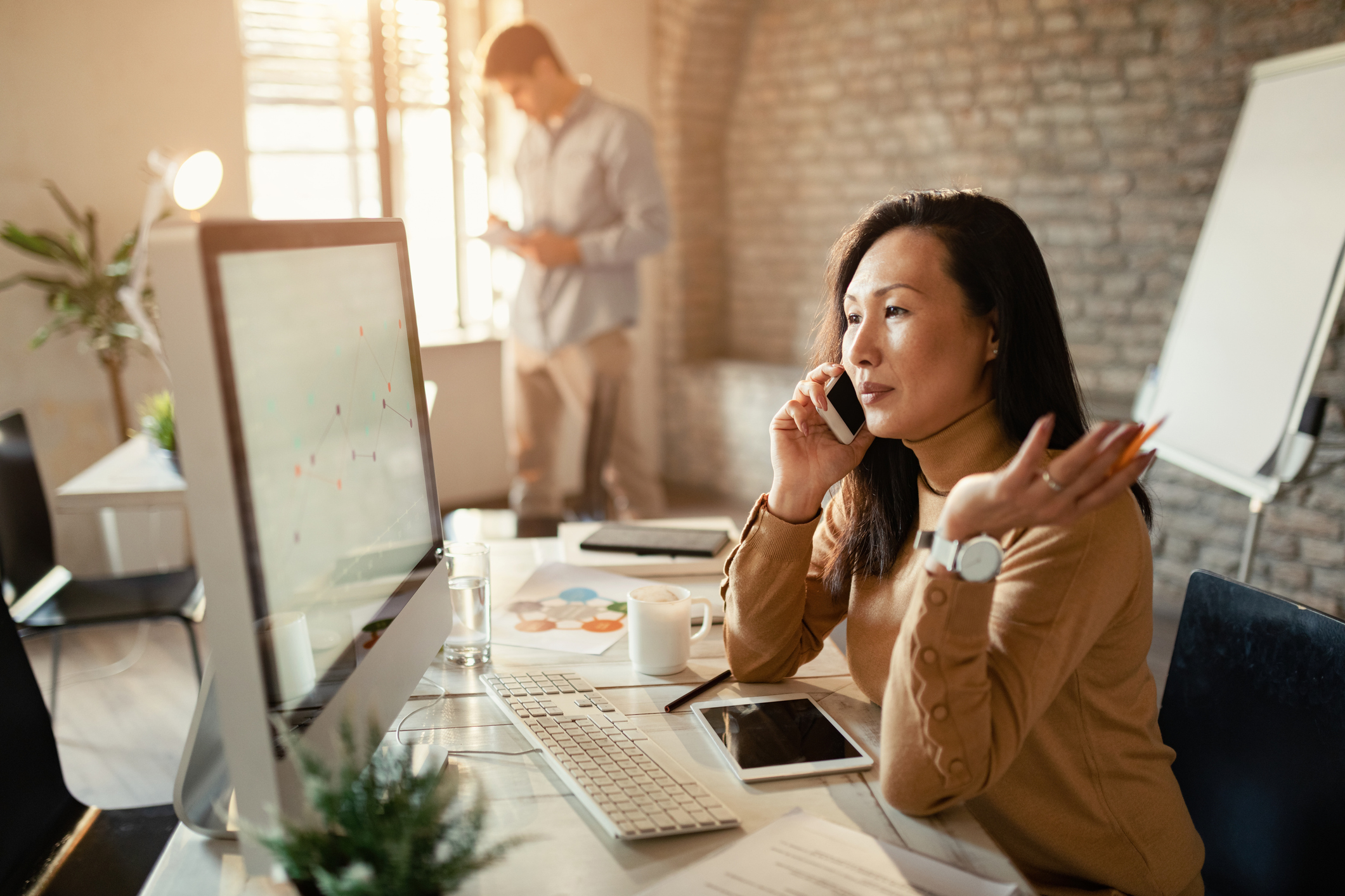 A businesswoman reviews charts on desktop PC while communicating on cell phone in the office for call-tracking blog.